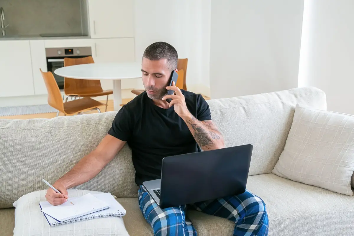 A man on a sofa holding a phone call while he writes notes in a notebook, a laptop on his lap