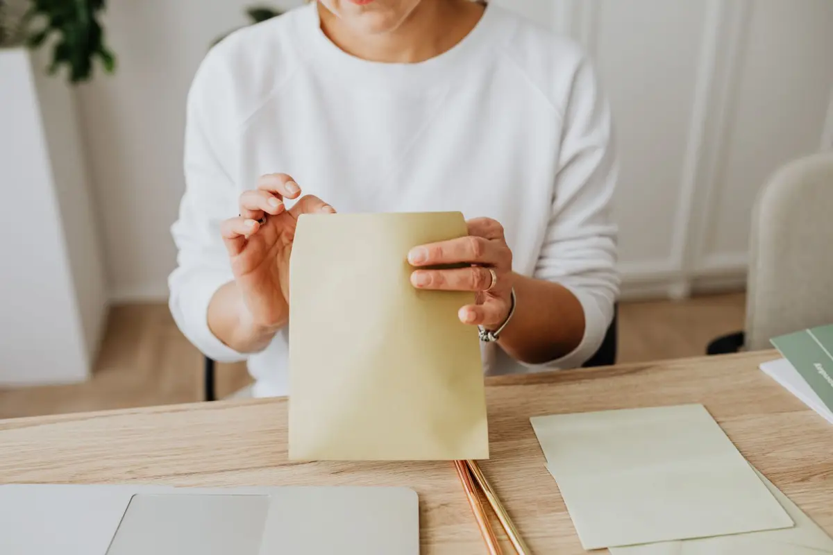 A woman at a wooden desk sealing a plain brown envelope, with blank stationery and a laptop in front of her