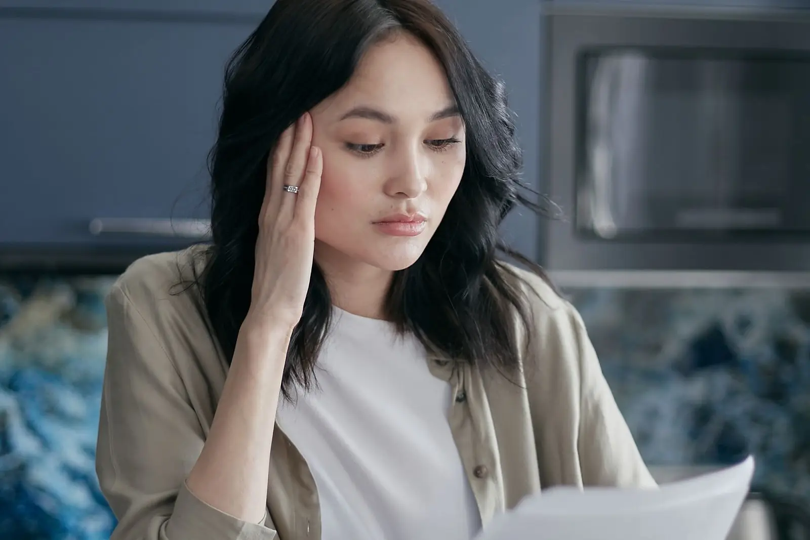 A woman at a kitchen table with a calculator and phone, frowning as she reads a printed letter