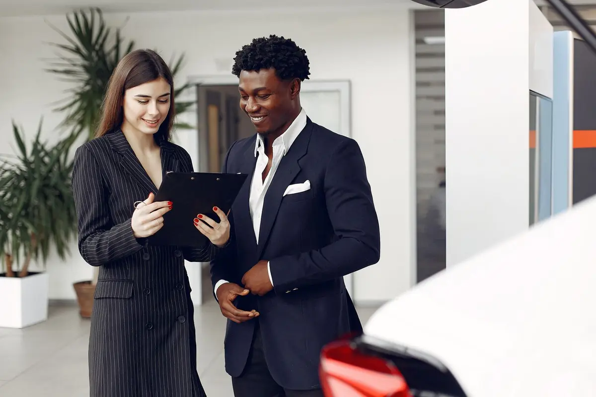 A car buyer in a suit and a dealership agent reading a document on a clipboard together in a bright showroom