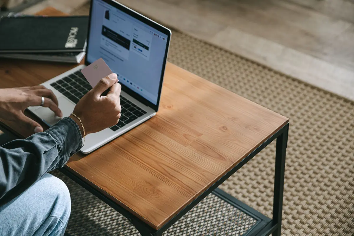 A person at a wooden coffee table holding a payment card in one hand while using a laptop with the other