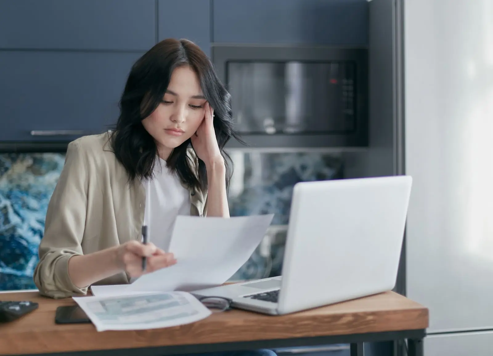 A woman at a kitchen table with a laptop, resting her head on her hand as she reads a printed page with a pen in hand