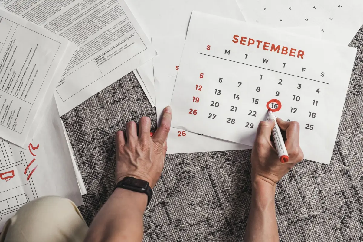 A hand circling a date in red marker on a paper calendar laid on a rug, with papers spread around it