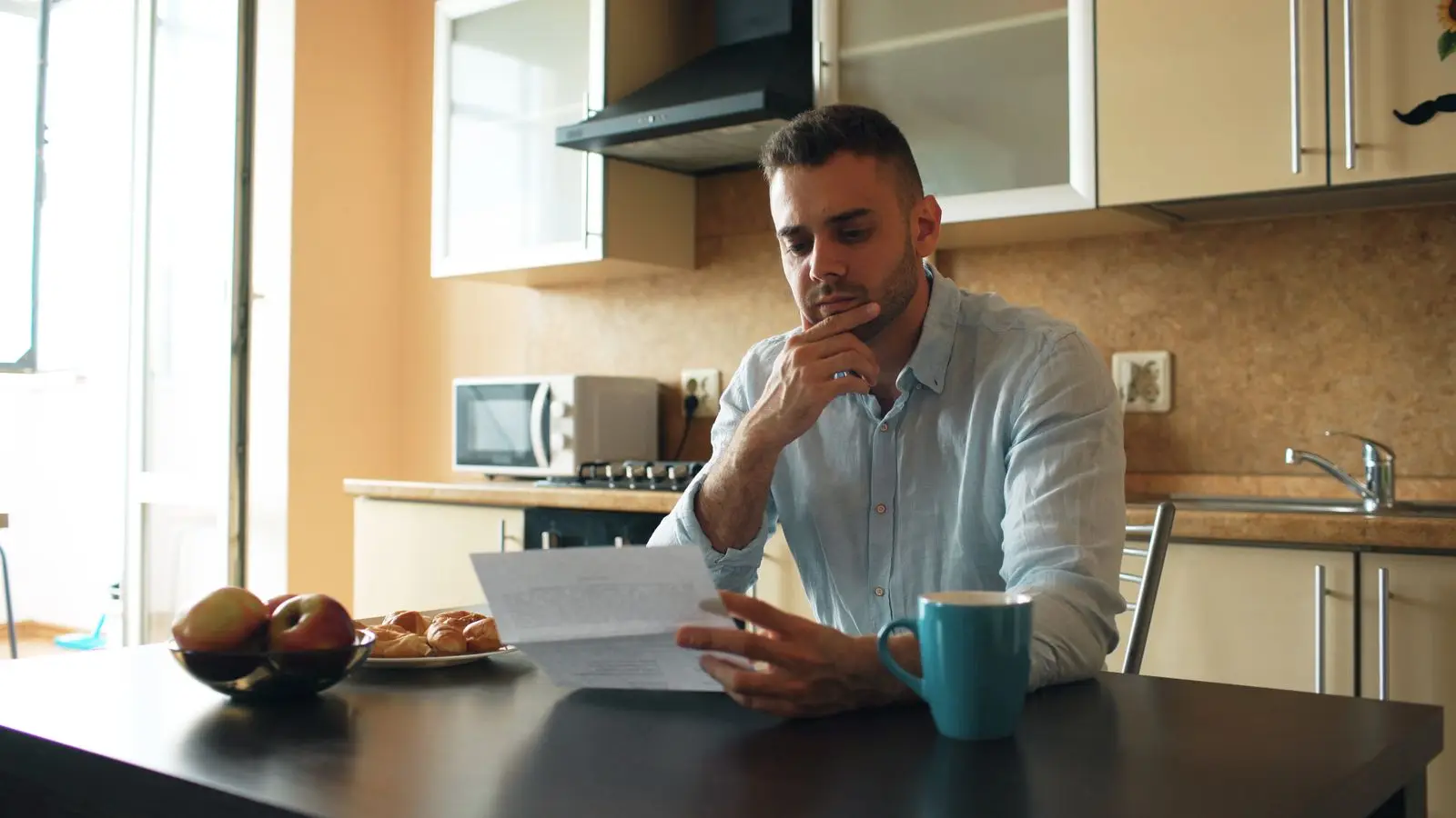 A man at his kitchen table, hand on his chin, reading a letter next to a cup of coffee