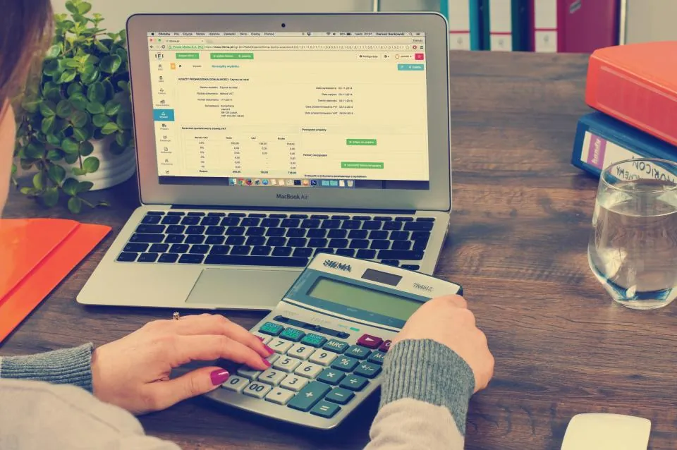 Hands entering numbers on a desktop calculator in front of a laptop showing an invoice in accounting software