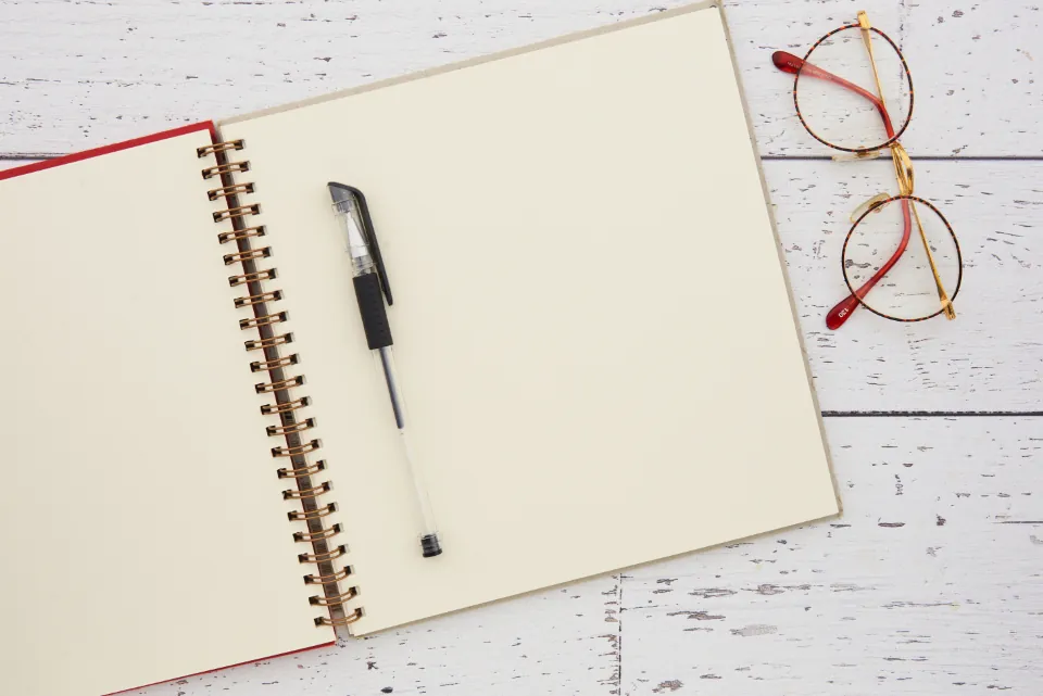 An open spiral notebook with blank pages and a black pen beside a pair of reading glasses on a white wooden table