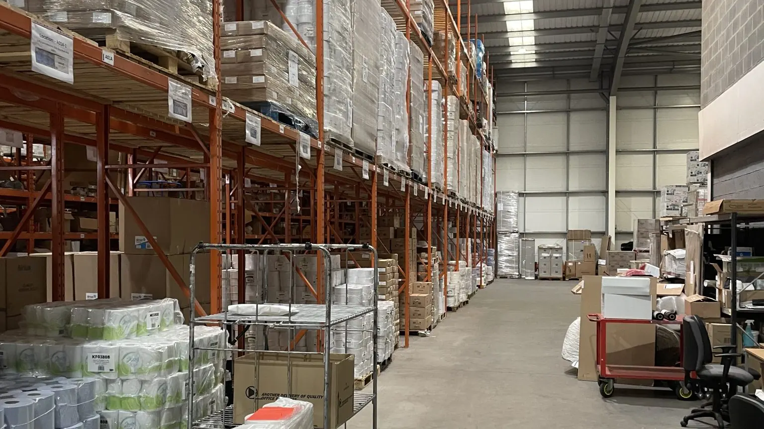 A long supply warehouse aisle with orange racks stacked high with shrink-wrapped pallets of boxes and paper goods